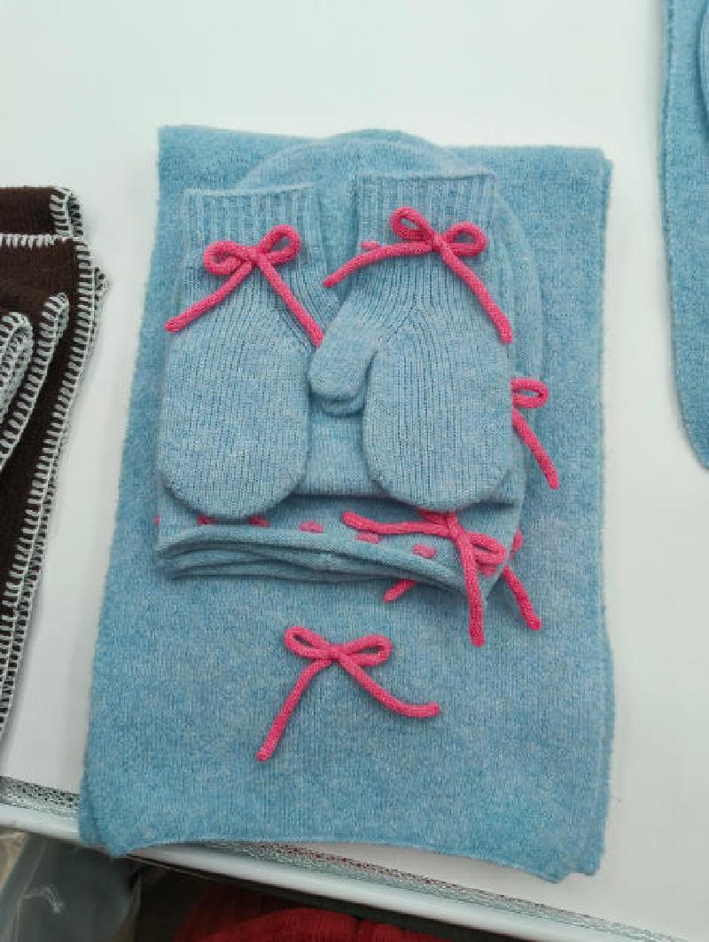 Baby blue knitted winter gift set including scarf, beanie hat and mittens with pink bow embellishments, displayed flat
