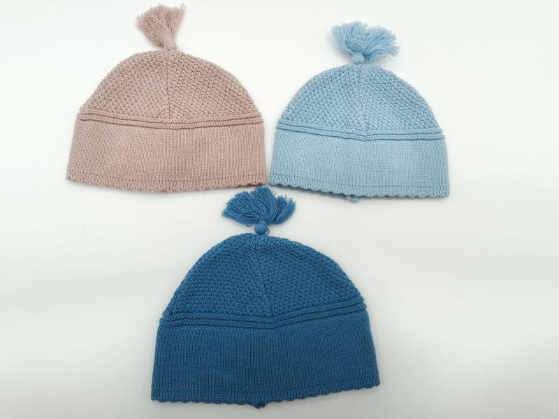 Three knitted baby beanies with tassels in dusty pink, light blue, and teal blue arranged on white background