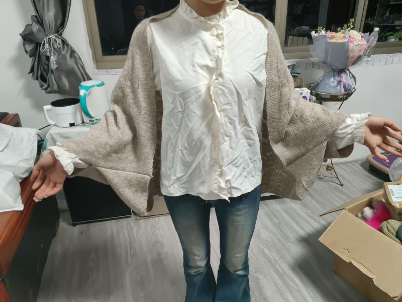 Woman wearing oversized beige knit cardigan layered over cream quilted ruffle-collar shirt with blue flare jeans indoors