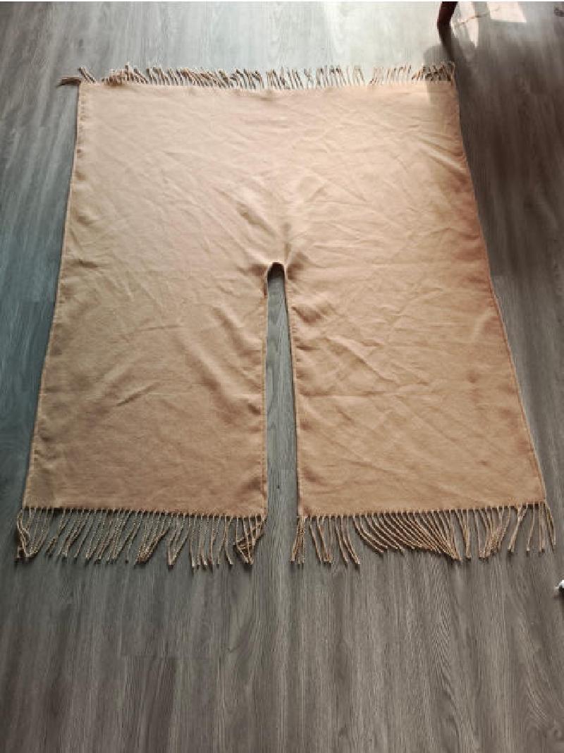 Flat lay of a beige woven poncho with center neck slit and twisted fringe along top and bottom edges on wood floor