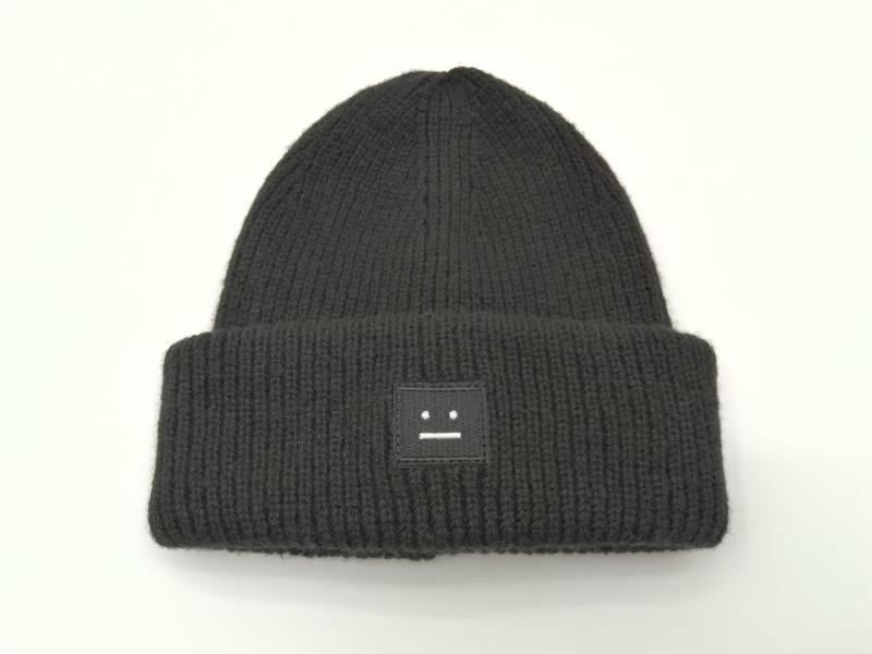 Black chunky ribbed knit beanie hat with folded cuff and small square embroidered neutral face logo patch on white background