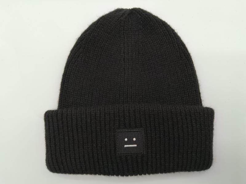 Black ribbed knit beanie hat with folded cuff and small embroidered face patch logo on white background