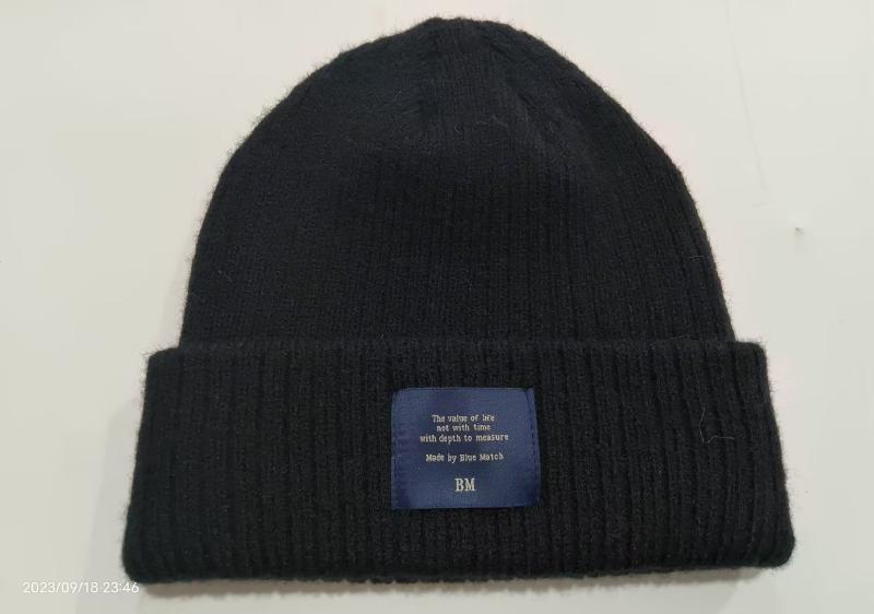 Black ribbed knit beanie hat with folded cuff and blue woven brand label patch on white background