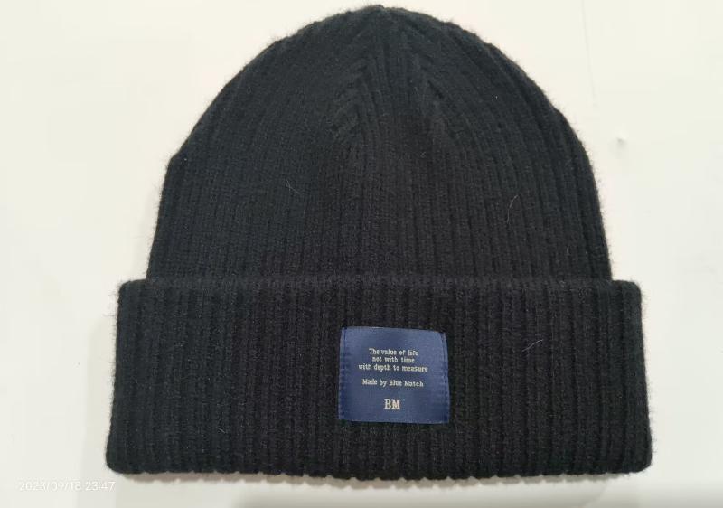 Black ribbed knit cuffed beanie with blue woven label patch on folded brim, soft textured wool winter hat