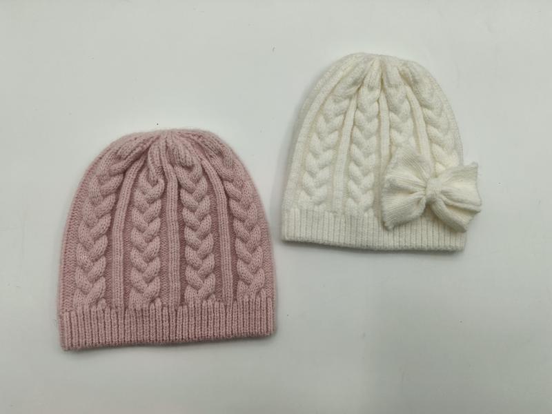 Two cable knit winter beanies side by side, one dusty pink and one cream white with a decorative bow on the side