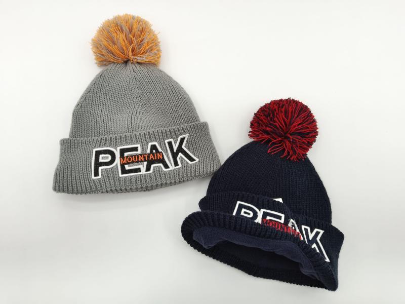 Two chunky knit pom-pom beanies in grey and navy blue with embroidered Peak Mountain logo, displayed on white background