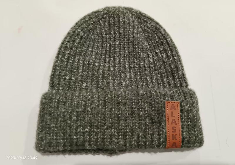 Olive green chunky ribbed knit beanie hat with folded cuff and brown leather Alaska patch label on white background