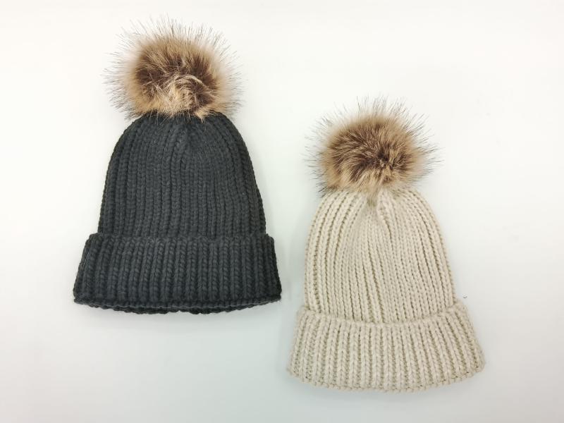 Two chunky ribbed knit beanies with faux fur pom poms in charcoal gray and cream beige on white background