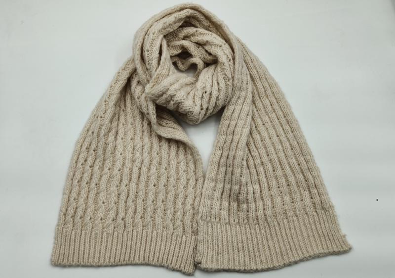 Cream beige cable knit scarf loosely coiled on white background showing textured pattern and ribbed hem ends