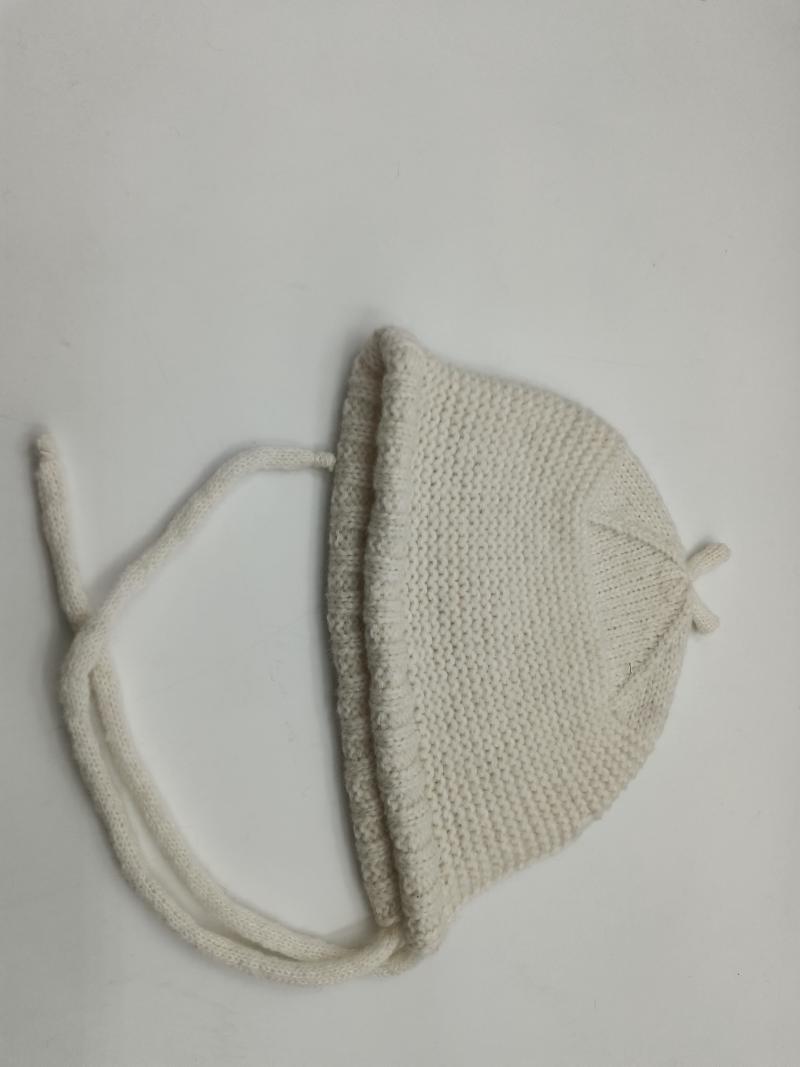 Cream ivory hand-knitted baby bonnet with chin tie strings laid flat on white surface, garter stitch texture