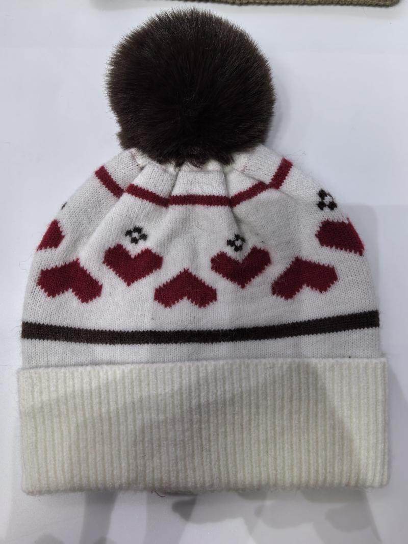 White knitted winter beanie hat with red heart pattern, dark brown faux fur pompom, and ribbed cuff on white background