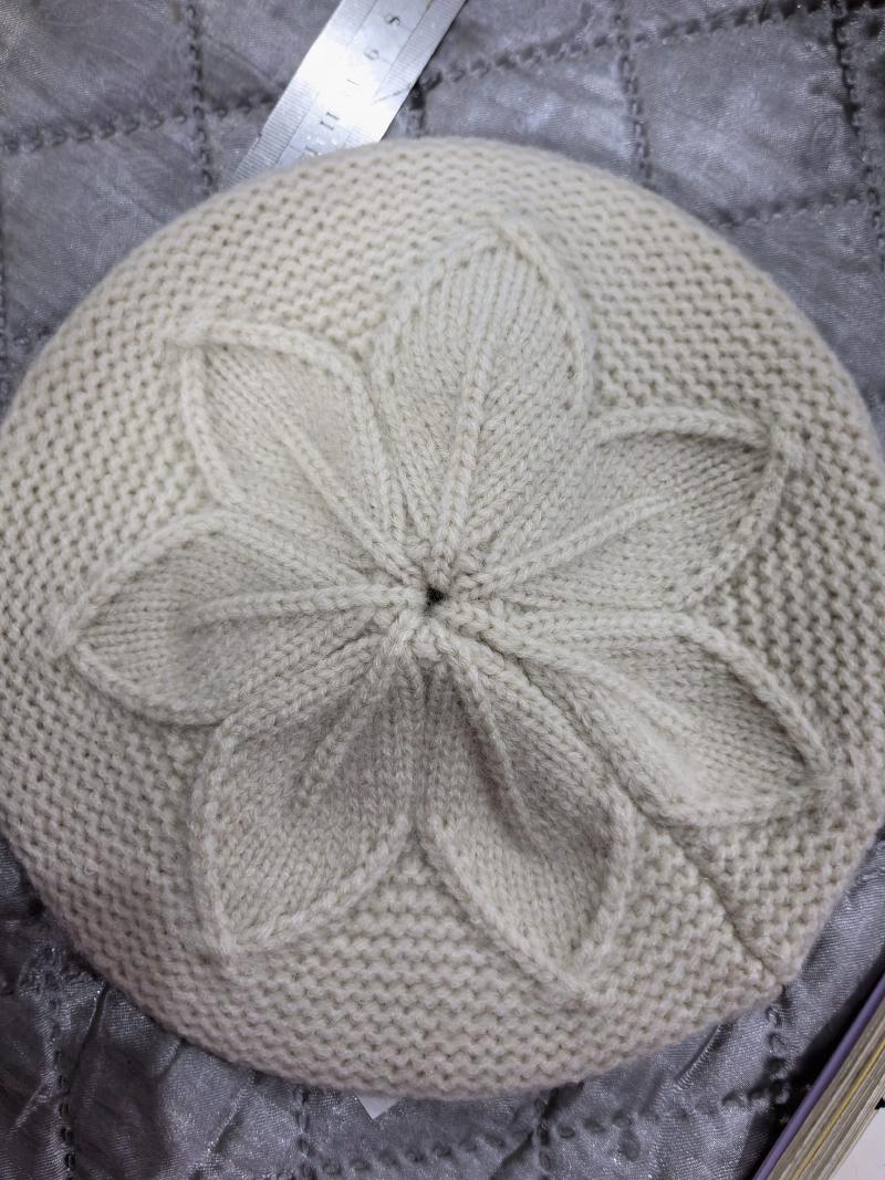 Top view of a cream knitted beret with a raised petal flower cable pattern at the crown, laid flat on gray quilted fabric