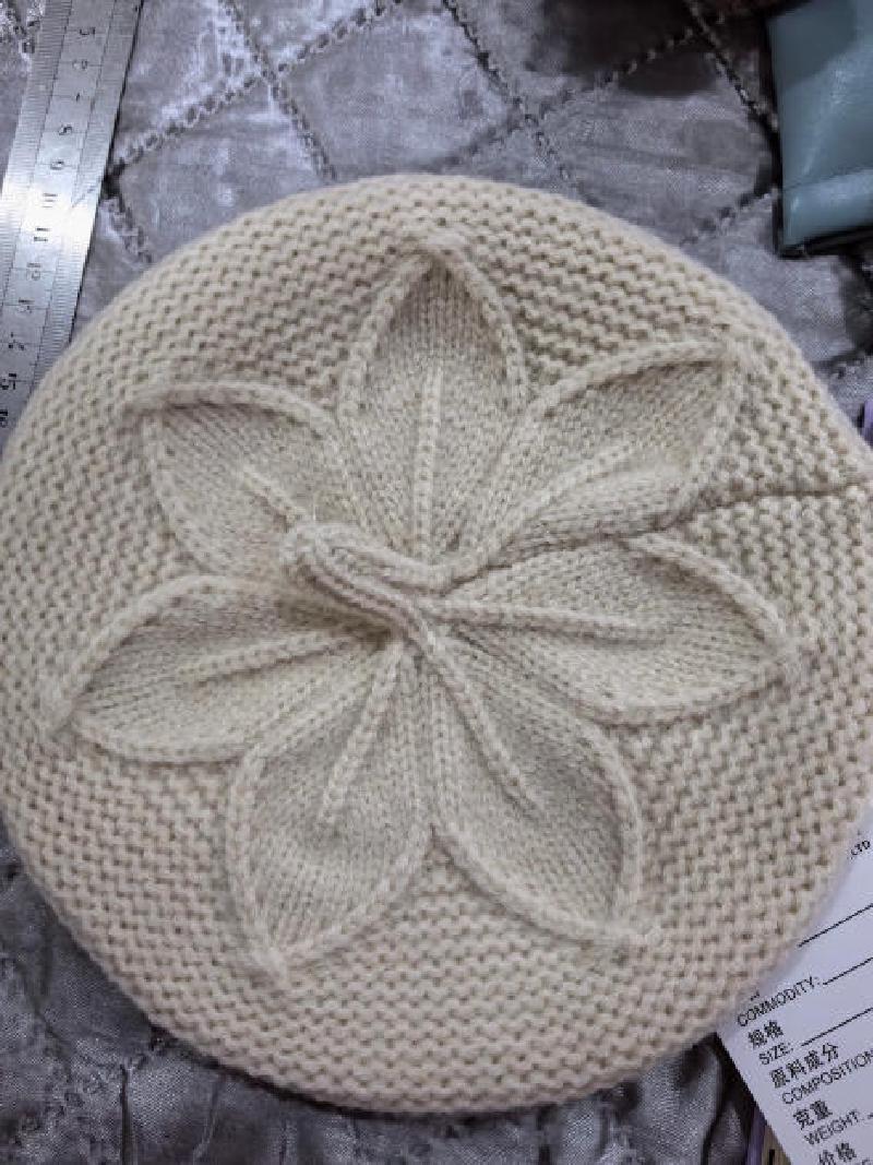 Cream knitted beret hat with raised floral leaf cable pattern at crown, laid flat on silver fabric