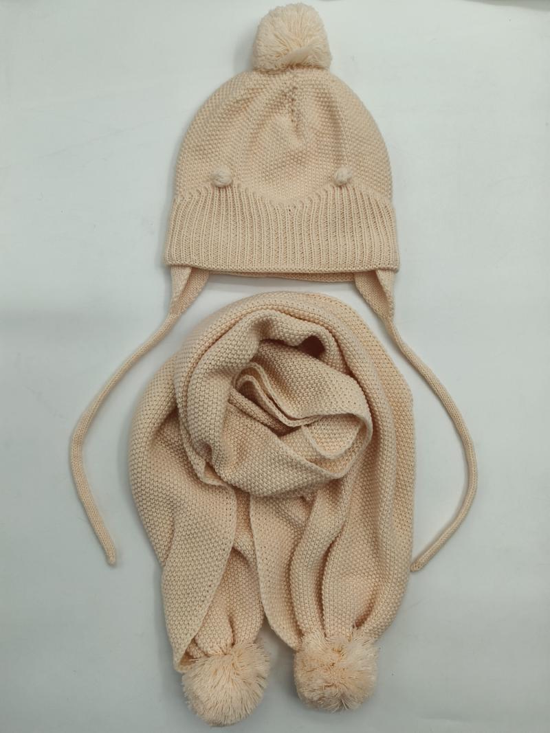 Cream knit children's winter hat with pom-pom top and ear flaps paired with matching knit scarf with pom-pom ends