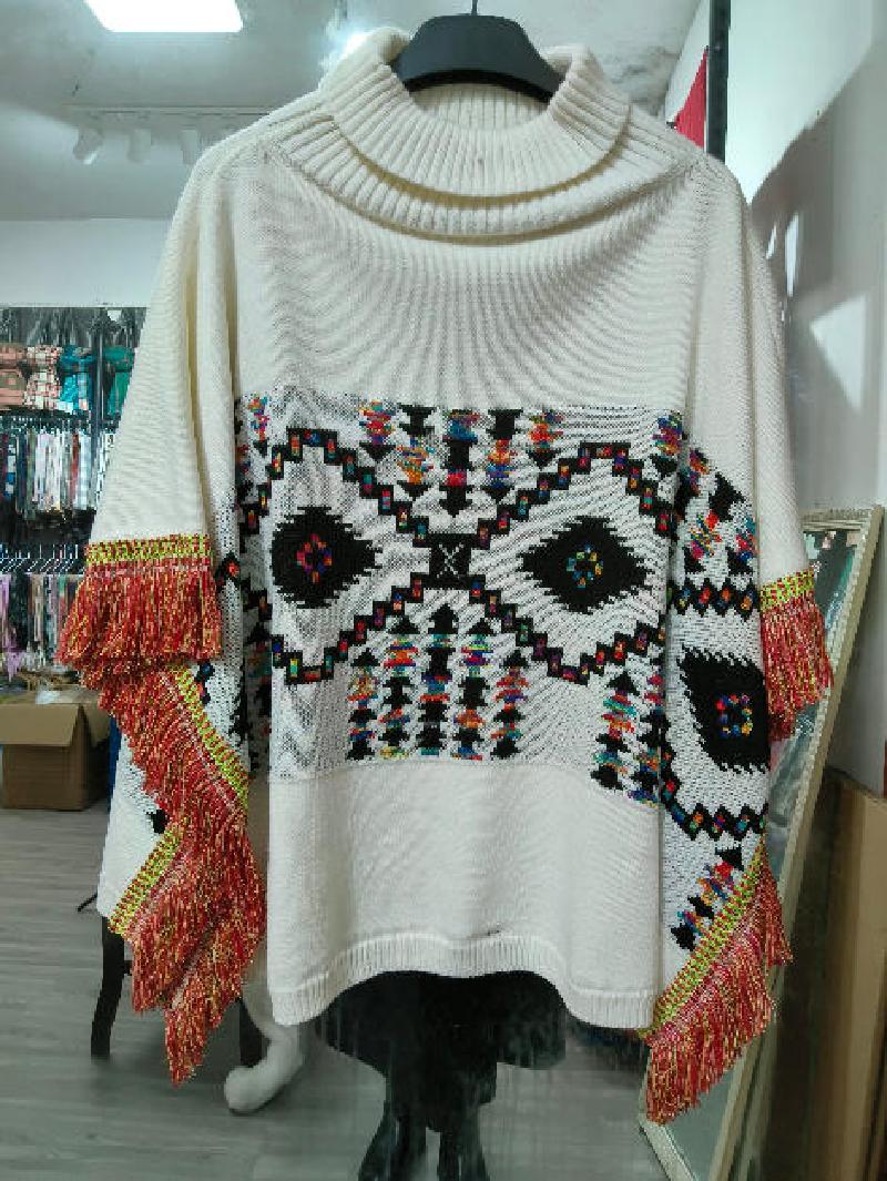 Cream oversized knit turtleneck poncho with multicolor Aztec geometric pattern and red fringe-trimmed sleeves on mannequin