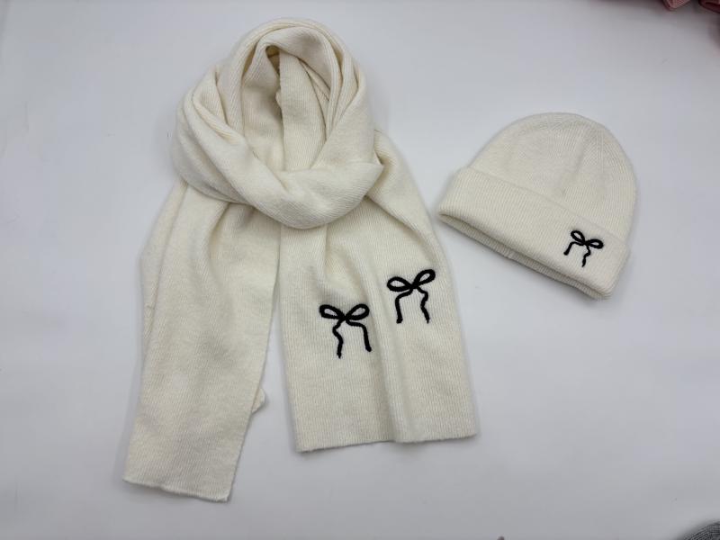 Cream ribbed knit winter hat and scarf set with black embroidered bow details laid flat on white surface