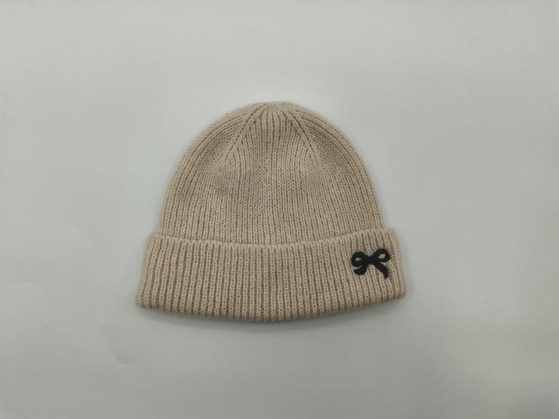 Cream ribbed knit beanie hat laid flat on white background featuring a small black embroidered bow on the folded cuff