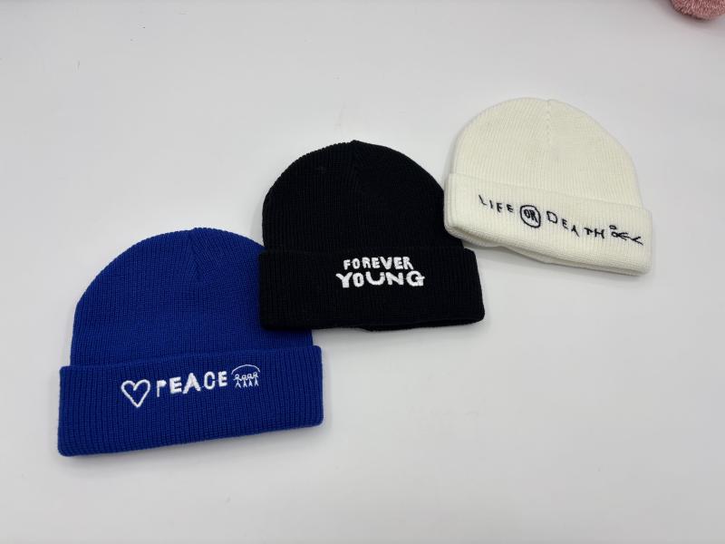 Three ribbed knit cuffed beanies in blue, black, and cream with embroidered slogans Peace, Forever Young, and Life or Death