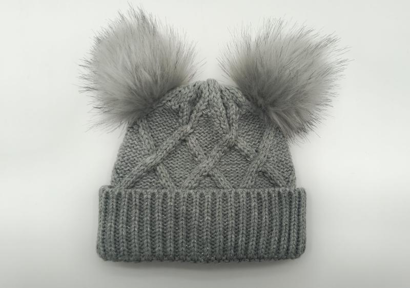 Gray cable knit winter beanie hat with two fluffy faux fur pom poms on top, ribbed cuff, flat lay on white background