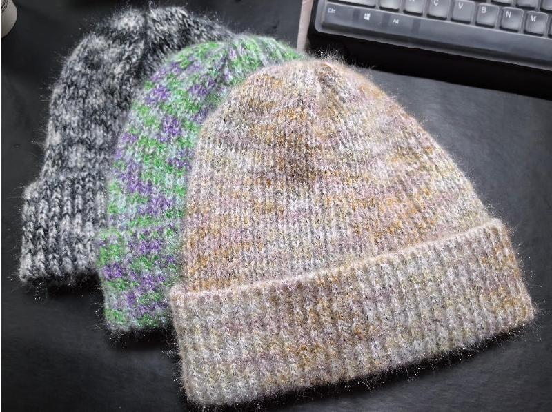 Three handknit wool blend beanie hats in gray marled, green purple stripes, and beige gold tones on dark surface