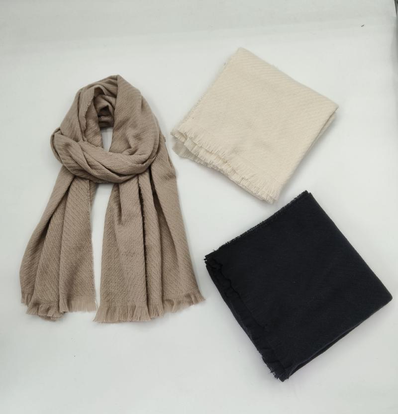 Three scarves displayed on white surface: draped taupe herringbone fringe scarf, folded cream scarf, and folded black scarf