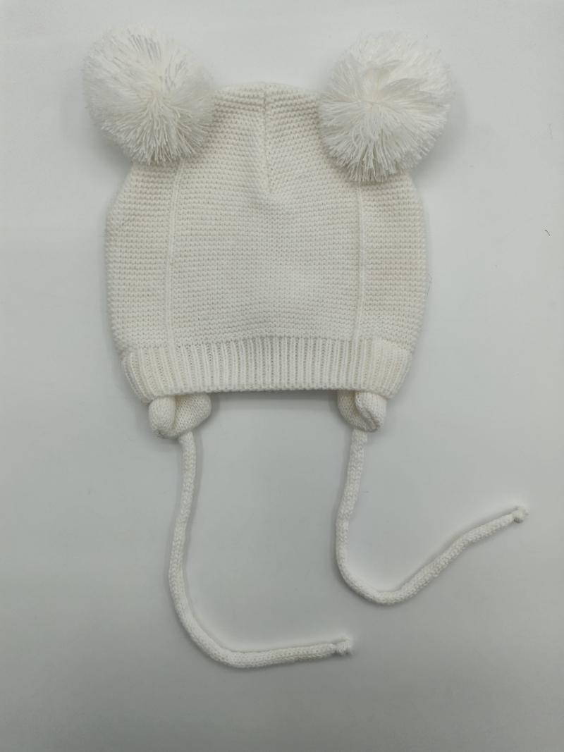 White knitted baby beanie with two fluffy pom poms on top and long chin tie strings, laid flat on white background