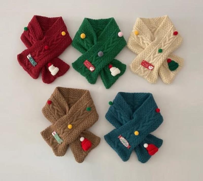 Five colorful children's cable knit crossover scarves in red, green, cream, brown, and teal with festive pom-pom and holiday charm decorations