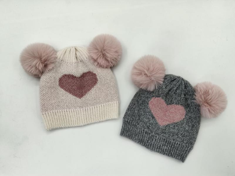 Two kids knit beanies with double faux fur pom poms and heart motif, one cream and one gray