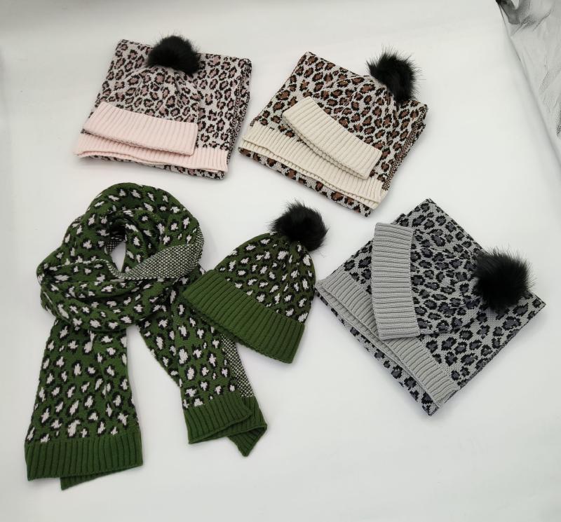 Four leopard print knitted hat and scarf sets in pink, cream, green, and gray with black faux fur pompoms displayed on white surface