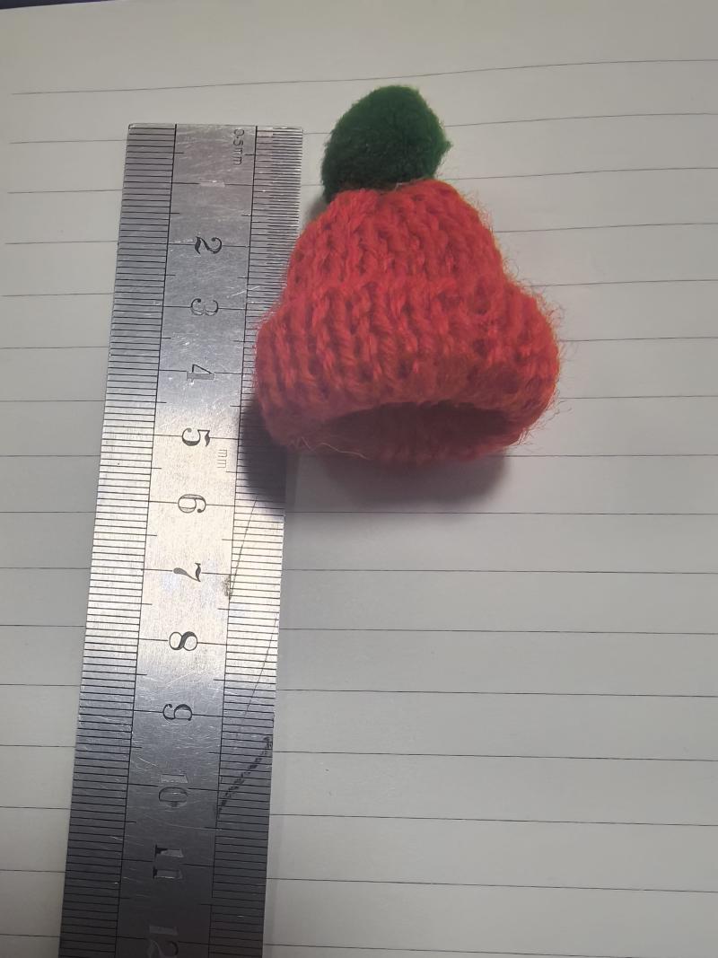 Tiny handknitted red wool beanie hat with green pom-pom measured against a ruler showing approximately 4cm height