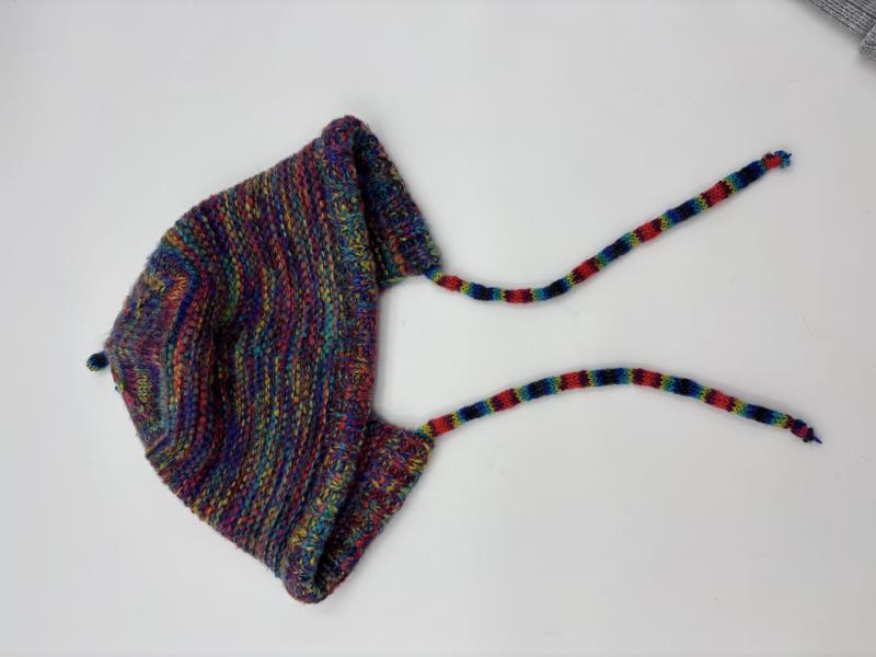 Handknit baby bonnet in multicolor variegated yarn with earflaps and braided tie strings, laid flat on white background